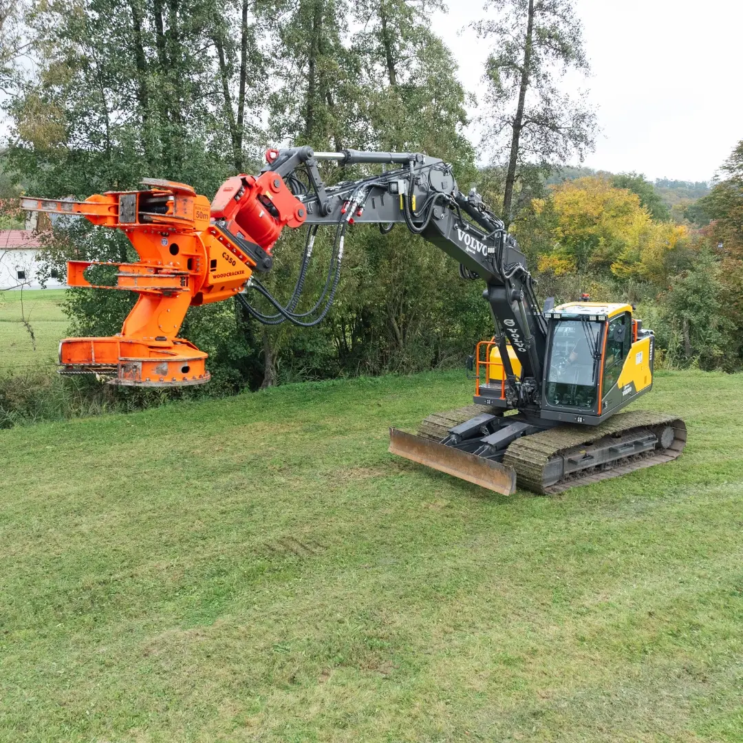 Moderner Volvo Forstbagger mit Holzspalter im Einsatz für professionelle Forstarbeiten in ländlicher Umgebung. Der Bagger mit seinem orangenen Greifarm ist ideal für schwere Erd- und Holzarbeiten geeignet. Perfekt für den Einsatz in der Forstwirtschaft zum Fällen und Zerkleinern von Bäumen. Einsatz in der Region Neustadt an der Aisch für effektive und schnelle Forst- und Erdarbeiten.
